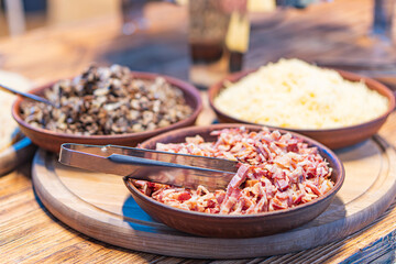 Image of a wooden tray with three dishes containing roasted nutsseeds, crispy bacon, and grated cheese possibly Parmesan Taken in an indoor setting with warm lighting, possibly during meal prepar