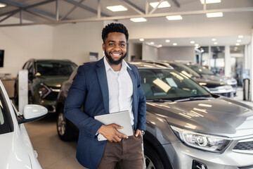 Portrait of smiling interracial car dealer standing at car showroom with tablet in hands.