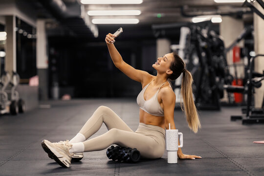 Smiling fit female athlete sitting on a gym floor and taking selfies.