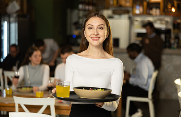 Girl waiter with tray walks between rows of tables. Employee of public catering cafe carries order to client, ready to serve guest of cafe establishment.