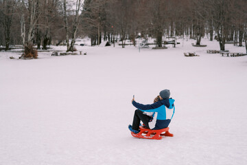 Dad taking smartphone selfie of himself and little kid sitting on sled on snowy lawn. Back view