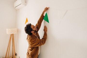 Man standing and laughing while hanging ribbon on wall, in apartment, preparing for birthday party