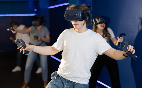 Excited modern young guy with gaming controllers in hands and VR goggles having fun with friends in virtual reality room. Concept of immersive interactive experience..