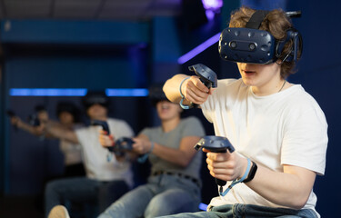 Excited modern young guy with gaming controllers in hands and VR goggles having fun with friends in...