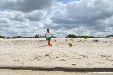 A woman dressed in green shorts, white shirt and dark glasses walks on the beach, next to a small dog, while kicking a ball during a cloudy day.