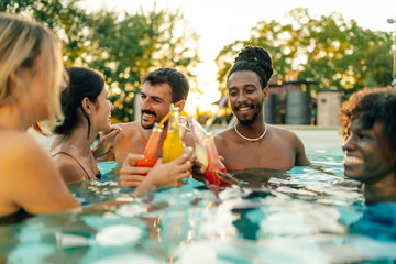 Friends toasting colorful drinks in swimming pool at sunset