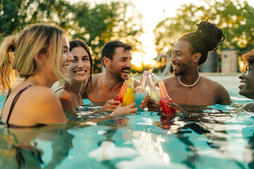 Friends toasting colorful drinks in swimming pool at sunset