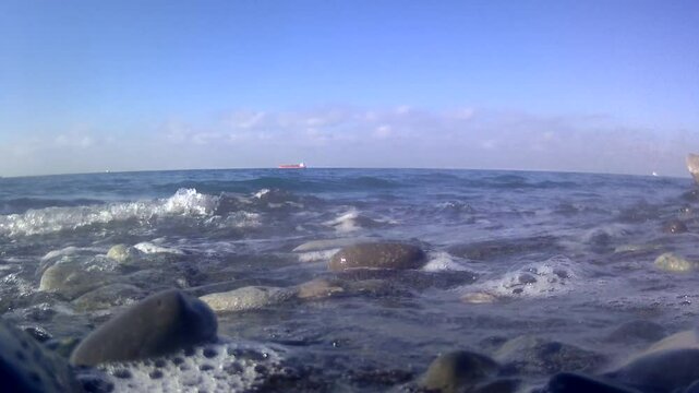 Close-up of the waves of the Black Sea washing the stone coast of Batumi. In the distance, a cargo ship can be seen on the horizon. The clear sky and calm sea surface create a peaceful atmosphere.
