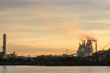 Obraz premium Silhouetted industrial factory set against a golden sunset, with lush greenery providing a serene contrast, captured in a peaceful forest setting, showcasing the juxtaposition of nature and industry