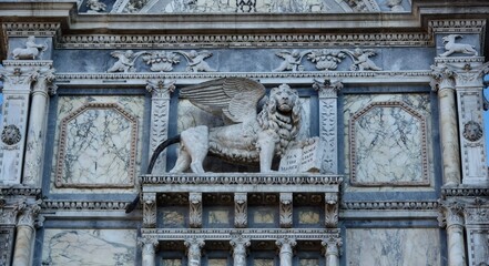 Obraz premium A detailed stone relief of the Winged Lion of St. Mark, a symbol of Venice, holding an open book with Latin text. The sculpture is set within an ornate marble and stone architectural facade.