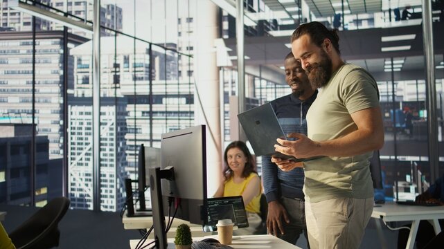 Cheerful admin holding laptop chatting with colleagues while working with machine learning tech. Man transforming data into predictive models using notebook and talking with coworkers, camera B