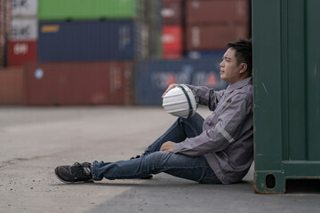 A tired logistics worker in a gray uniform sits on the ground at a busy shipping container yard, holding his safety helmet while resting after a long shift in the freight transportation industry.