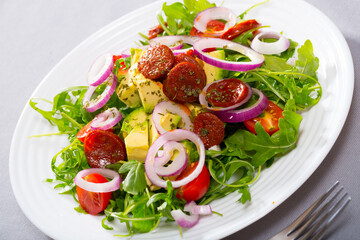 Delicious salad of arugula with chopped ripe avocado, roasted chorizo sausage, tomatoes and purple onion