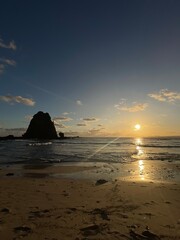 landscape view of Papuma Beach in Jember, featuring a rocky islet standing tall against the warm hues of the setting sun. The sky transitions from deep blue to orange. Papuma Beach Jember, Indonesia