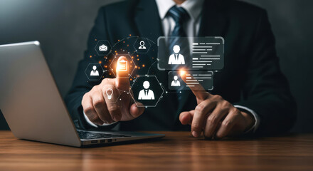 A business professional in a suit engages with a digital display of icons representing communication and data management tools while using a laptop. The setting reflects a modern work environment