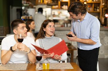 During communicating with couple customers, man waiter writes down order of dishes and drinks, answers customers questions about offer from chef. Friends in restaurant makes order to waiter