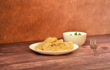 A plate of freshly cooked dumplings, a fork and a cup of sour cream with a sprig of parsley on a wooden table.