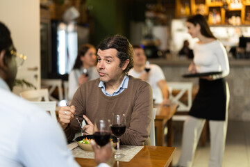 Man in cafe sits opposite African male companion, has nice conversation and discusses news of day. Cozy atmosphere of cafe, meeting place for friends, business partners, colleagues.
