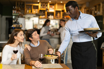 African waiter brings cooked food from the chef for a couple in a restaurant. Restaurant customers drink European wine while waiting for food. Husband and wife order food from the waiter