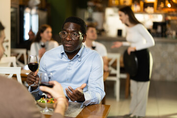 African man in cafe sits opposite male companion, has nice conversation and discusses news of day. Cozy atmosphere of cafe, meeting place for friends, business partners, colleagues.