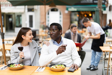 European woman and African husband are sitting on the terrace of a restaurant in warm clothes and happily chatting. International couple is actively talking, discussing delicious food and drinking