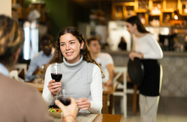Smiling woman in warm clothes sitting with a glass of red wine and talking to man in a cozy restaurant. Romantic dinner for a couple in a European restaurant