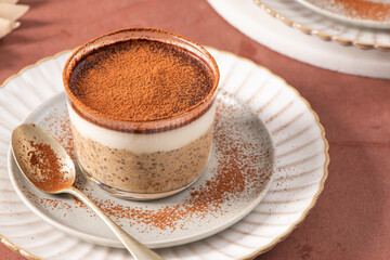 Tiramisu chia pudding in a glass, topped with cocoa powder, served on a white plate with a spoon,...