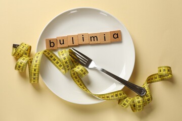 Plate with word Bulimia, fork and measuring tape on beige background, top view
