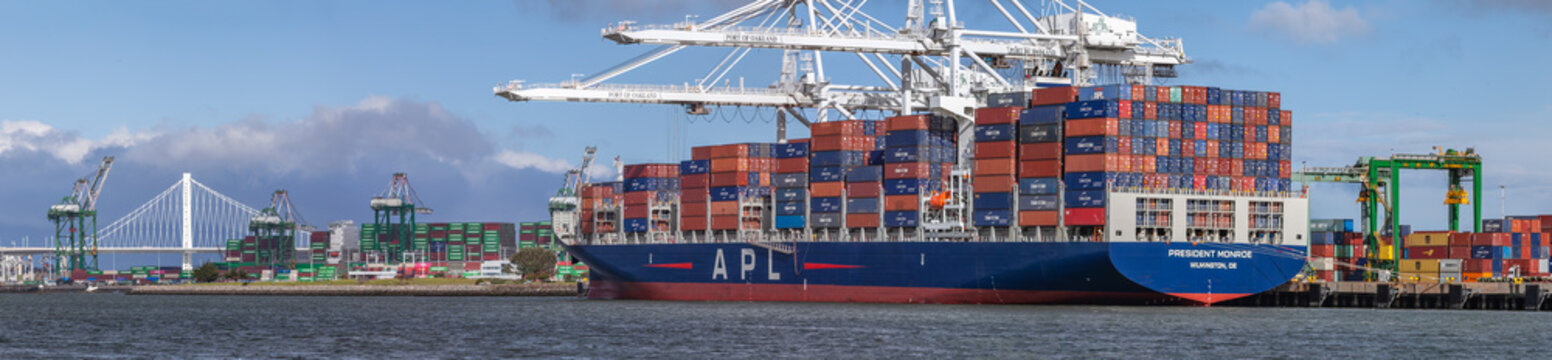 Large cargo ship docked at the Port of Oakland with colorful shipping containers and industrial cranes under an overcast sky, with the name APL on the side