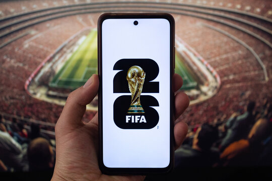 A male hand holding a smartphone with fifa world cup 2026 logo over white background and blurred football stadium at background