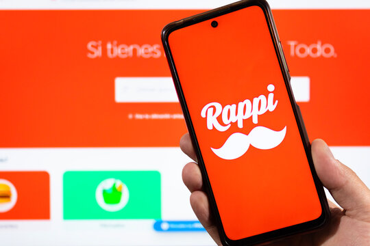 A male hand holding a smartphone with his right hand and Rappi orange logo and blurred rappi interface at background