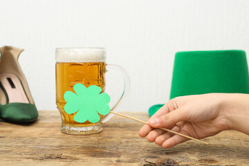 Female hand with paper clover, mug of beer and leprechaun hat on wooden table against white...