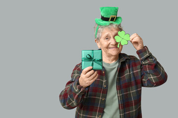 Senior woman in leprechaun hat with clover and gift box on grey background. St. Patrick's Day...