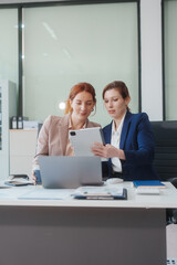 Two Caucasian women work in an office, happily using laptops and computers at a desk, discussing...