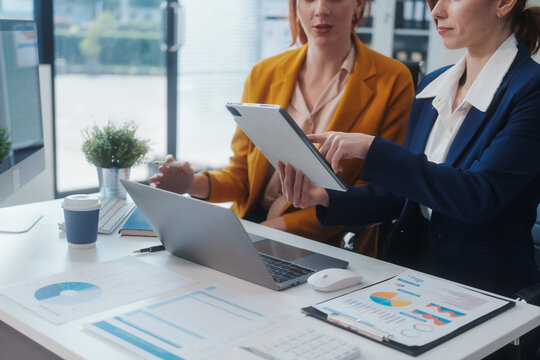 Two Caucasian women work in an office, happily using laptops and computers at a desk, discussing financial projects, analyzing financial statements,collaborating in a meeting to ensure effective