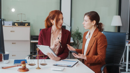 Two Caucasian women meet at a table, discussing legal matters. As lawyers, they advise clients on...