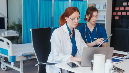 Two Caucasian female doctors work at a desk, meeting and discussing patient treatment while using a...