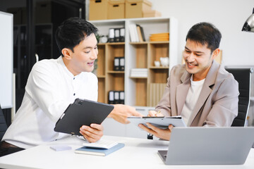 Fototapeta premium Two Asian businessmen sit in the office using laptops. They manage business tasks, communicate with clients, analyze data, and oversee projects to ensure success and efficiency in their operations