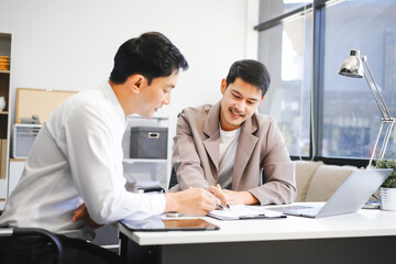 Two Asian businessmen sit in the office using laptops. They manage business tasks, communicate with clients, analyze data, and oversee projects to ensure success and efficiency in their operations