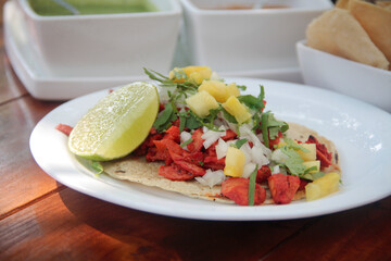 Authentic taco al pastor on a warm corn tortilla, featuring thinly sliced marinated pork, tangy pickled pineapple, fresh cilantro, and diced white onion on tropical beach beach club setting.