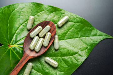 Pile of herbal capsules on wooden spoon with herbs and green natural Betel pepper leaf background