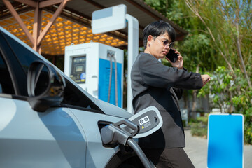 Person charging electric vehicle while making a phone call in a modern station during daytime