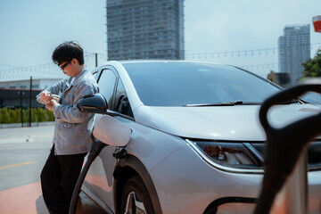 Naklejka premium Young man charging electric vehicle while enjoying coffee at sunny outdoor location