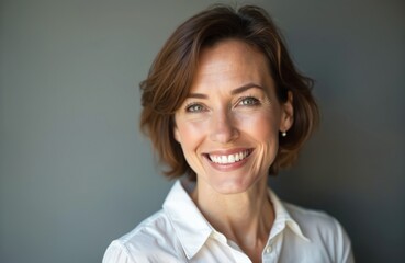 Headshot of cheerful middle aged woman smiling in studio. Mature model with short brown hair wears classic white shirt. Natural makeup, beautiful smile. Represents beauty, confidence, healthy aging.