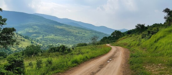 Fototapeta premium Winding dirt road in lush green countryside surrounded by mountains under a cloudy sky with patches of sunlight illuminating the landscape