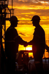Fototapeta premium Silhouettes of two workers shaking hands in front of oil pumps against a sunset sky, featuring the silhouette of an industrial area with machinery and pump jacks during dusk.