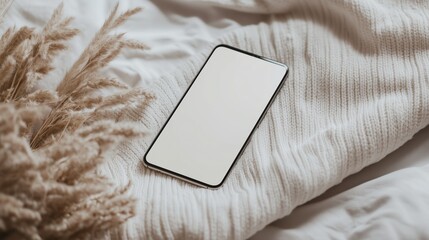Smartphone with a blank white screen resting on a cozy knitted fabric with dried pampas grass. Warm and aesthetic digital concept.
