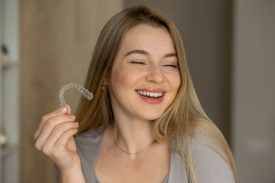 Smiling woman holding clear dental aligner in home interior