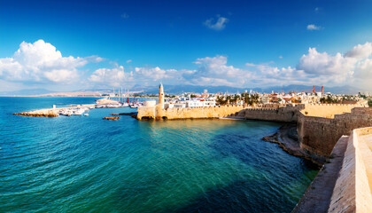 Ancient City by the Sea: A panorama captures the sun-drenched old city, its fortified walls and buildings rising from the edge of the vibrant blue sea, framed by a sky dotted with fluffy clouds. 