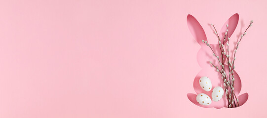 Elegant silhouette of Easter bunny filled with blossom pussy willow branches and painted Easter eggs on light pink background. Flat lay. Banner.